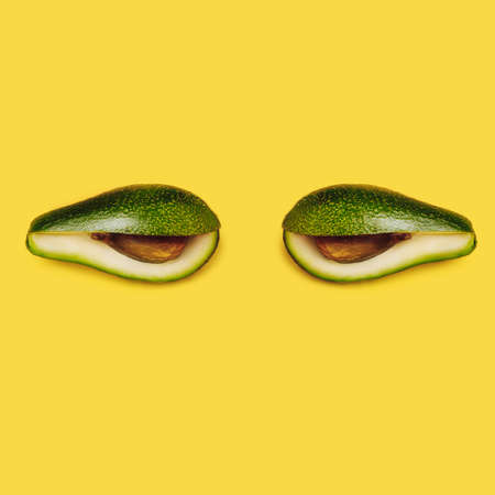 Fresh cut avocado shaped like eyes isolated on vibrant yellow background. Creative surreal food concept. Patches under the eyes morning care facial mask. Flat lay.の写真素材