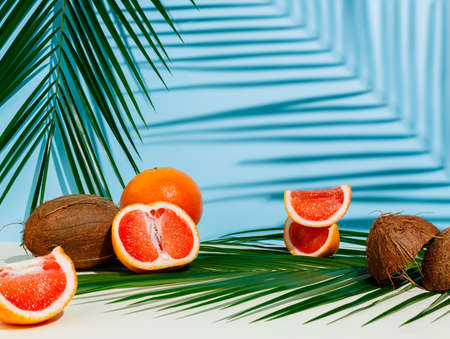 Natural summer scene with fresh grapefruits cut into slices and coconut placed on a palm leaf with shadows on blue background. Creative food concept. Assortment of tropical, exotic fruit.の写真素材