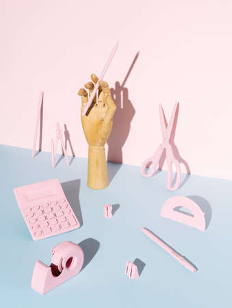 Artificial hand holding a pencil with office equipment on two tone pastel blue and pink background....