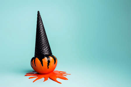 Black ice cream cone as a witch hat on top of orange pumpkin with black dripping paint isolated on a pastel blue background. Creative Halloween concept. Spooky and scary October party. Fall vegetable.の写真素材