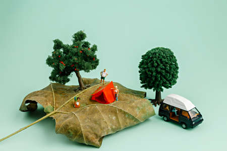 Camping in nature on a dry autumn leaf with a small tent, miniature people, two trees and a camping van on pastel green background. Creative camping or fall outdoor activity concept.の写真素材