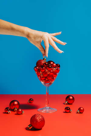 A woman's hand picking a Xmas bauble from the martini cocktail glass filled with red Christmas tree balls places on the red table with a blue background. Creative Xmas or New year celebration concept.の写真素材