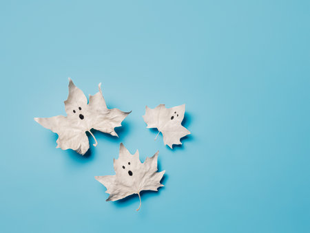 Ghosts made with white maple leaves on a bright blue background with copy space. Creative Halloween party creative concept. Minimal invitation card aesthetic. Cute and spooky visual trend.の写真素材