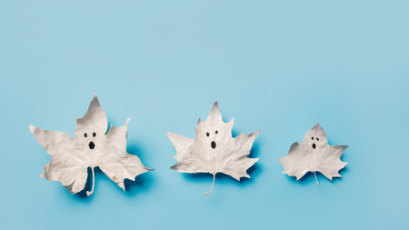 Three white maple leaves looking like ghosts on a bright blue background with copy space. Creative Halloween party creative concept. Minimal invitation card aesthetic. Cute and spooky visual trend.の写真素材
