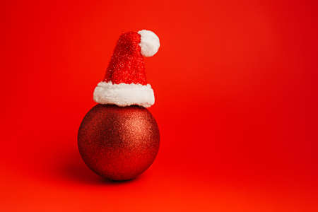 Creative Christmas layout made with shiny bauble ornament with Santa Claus cap on vibrant red background. Minimal Xmas or New Year celebration concept. Winter festive composition with copy space.の写真素材