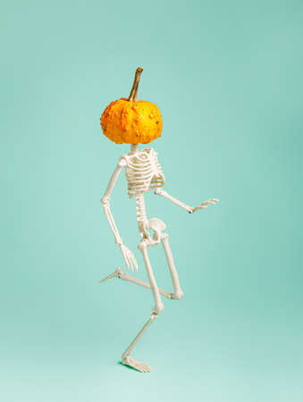 Human skeleton with orange pumpkin head dancing and waling against pastel green background. Creative Halloween concept. Minimal spooky or scary idea. 31th October party invitation card.の写真素材