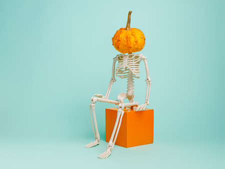 Human skeleton with orange pumpkin head sitting on the cube on pastel green background. Creative...