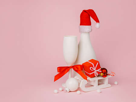 White champagne bottle with Santa's hat, white glass and ornament baubles with sleigh on pastel pink background. Creative New Year party celebration concept. Minimal drink or toast composition.の写真素材