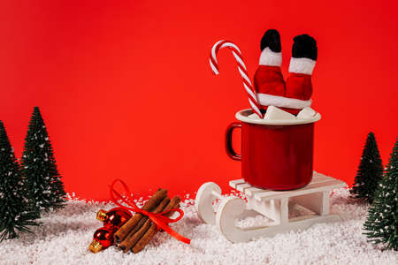 Christmas scene with Santa Claus stuck in red mug of hot drink with snow and pine tree forest on blue red background. Creative Xmas or New Year holiday concept. Winter traditional funny banner idea.の写真素材