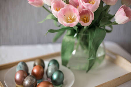 Beautiful colored shiny easter eggs on the white plate. Pink tulips flowers bouquet. Happy easter concept.の写真素材