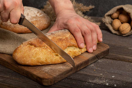 Male hands with a bread knife cut fresh crispy baguette on a wooden board. Sliced traditional french...