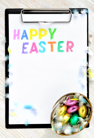 A tablet for paper with an inscription on a white Merry Christmas leaf and a small basket with chocolate eggs in colorful wrappingの写真素材