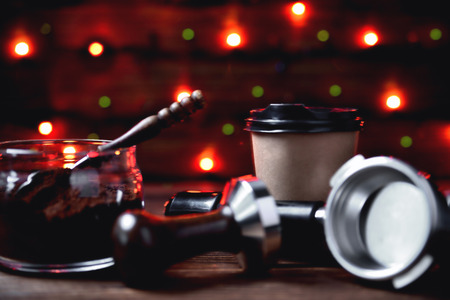 temper portofilter glass jar with ground coffee wooden coffee spoon paper cup on a natural wooden brown background with colored lights blurred foreground close-up copy spaceの写真素材