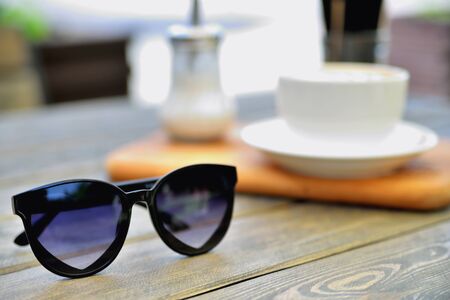 cup of coffee on a wooden stand is on the table in a cafe summer morning, sunglasses in the foreground, close-upの写真素材