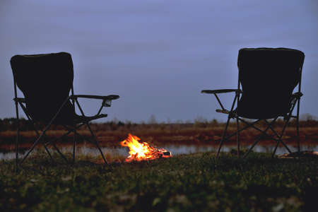 A pair of tourist chairs, a folding chair, a bonfire on the river bank in the late evening, twilight in the fall at sunset, a big bonfire in the dark, relaxation and rest, the beginning of autumn, tourism and travel, a close -up of a copy spaceの写真素材