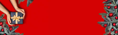 Web-banner with gift box in female hands and fir branches on red background. Flat lay, top view, place for text.の写真素材