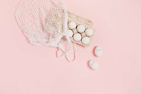 Happy Easter. White eggs with painted eyes in paper packaging and white mesh bag on pink background. Flat lay, top view, copy space.の写真素材