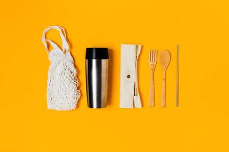 A group of eco friendly kitchen accessories. Thermo mug, bamboo cutlery, reusable drinking tube on yellow background.の写真素材