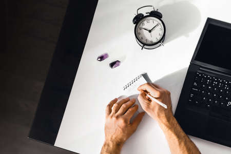 The man working at home on the remote. A black-and-white workplace with a laptop, alarm clock, notepad and men's hands.の写真素材