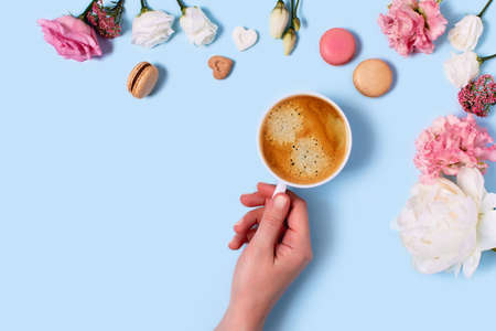Blue background with macaroons and a Cup of coffee in a hand surrounded by peonies and roses. Top...