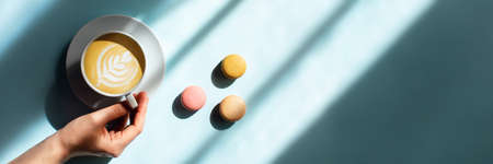 Blue background with cup of tasty coffee in a woman hand and color macaroons in sunlight.の写真素材