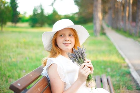 red-haired girl in a white hat sits on a park bench with a bouquet of lavender.の写真素材