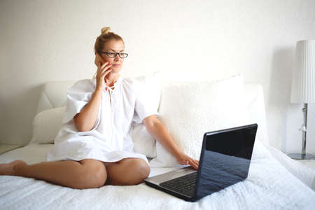 young woman on a white bed works for a laptop. remote work concept.の写真素材