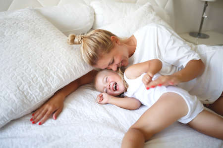 young woman hugging with her daughter on a white bed.の写真素材