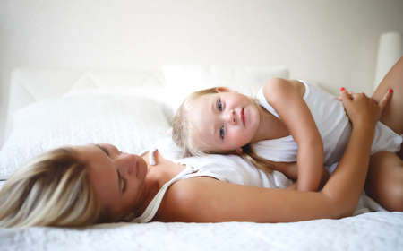 young woman hugs with her daughter on a white bedの写真素材