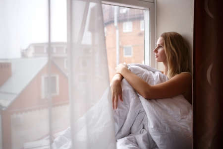 young woman wrapped in a blanket sits on the windowsill by a large window.の写真素材