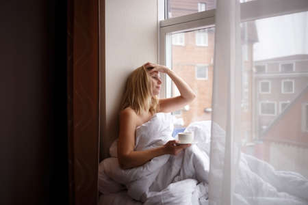 young woman wrapped in a blanket sits on the windowsill by a large window.の写真素材