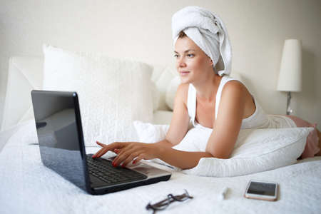 young woman on a white bed works for a laptop. remote work concept.の写真素材