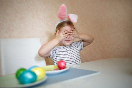 little cute blue-eyed girl in bunny ears paints Easter eggs.