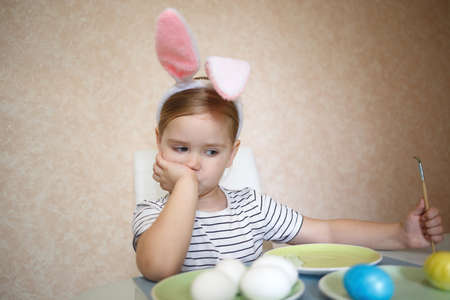 little cute blue-eyed girl in bunny ears paints Easter eggs.