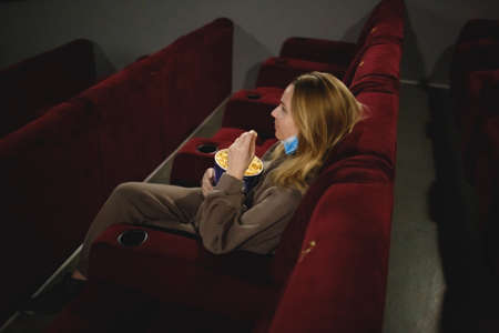 young woman in medical mask eating popcorn sitting in the cinema