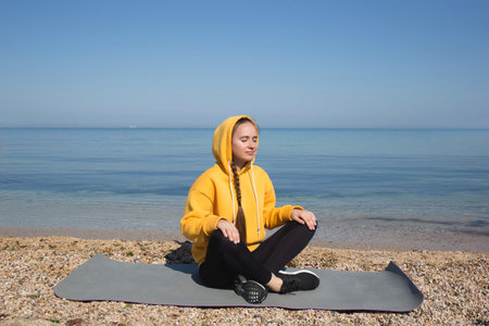 young woman meditates while sitting on yoga mat outdoors by the sea.