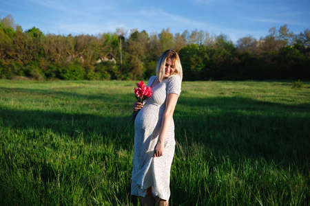 young pregnant woman on a green meadow walking with a bouquet of flowers.の写真素材