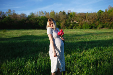 young pregnant woman on a green meadow walking with a bouquet of flowers.の写真素材