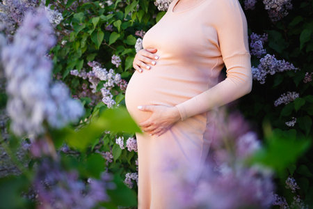 young pregnant woman tenderly hugs her belly surrounded by lilac flowers.