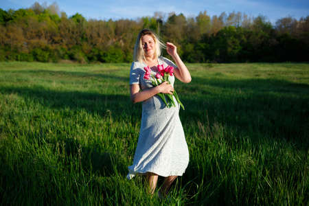 young pregnant woman on a green meadow walking with a bouquet of flowers.の写真素材