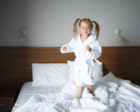 little cute girl in a white robe jumping on the bed.の写真素材