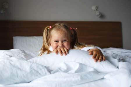 portrait of a little cute girl in a white robe lying on the bed in the bedroom.の写真素材