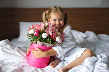 Little cute girl in a white robe holds a box of flowers while sitting on the bed.の写真素材