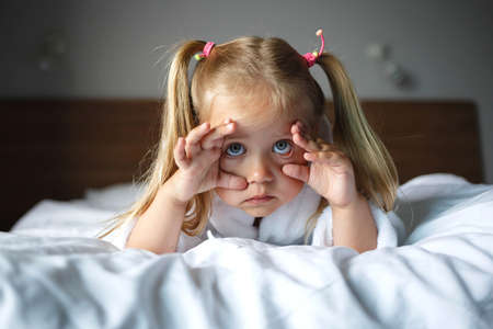 portrait of a little cute girl in a white robe lying on the bed in the bedroom.の写真素材