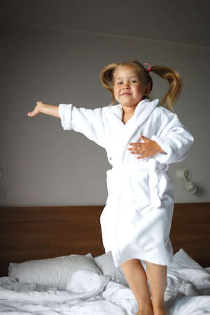 little cute girl in a white robe jumping on the bed.の写真素材