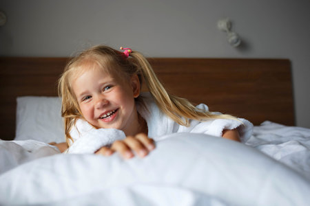 portrait of a little cute girl in a white robe lying on the bed in the bedroom.の写真素材