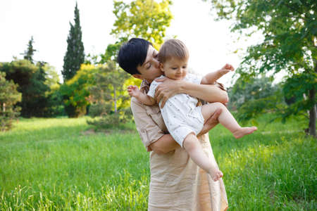 young mom kisses her little son while sitting in the park on the lawn.