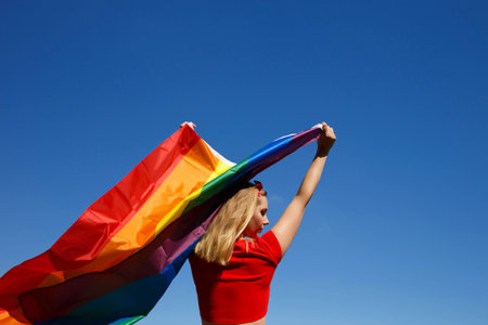 girl with lgbt flag behind her back against a bright blue sky.の写真素材