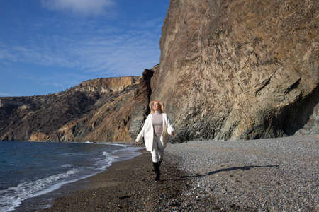 a young beautiful woman runs along a pebble beach against the backdrop of rocks and the sea.の写真素材