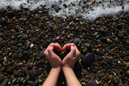 female hands with a beautiful manicure depict a heart on the background of wet pebbles on the beach.の写真素材